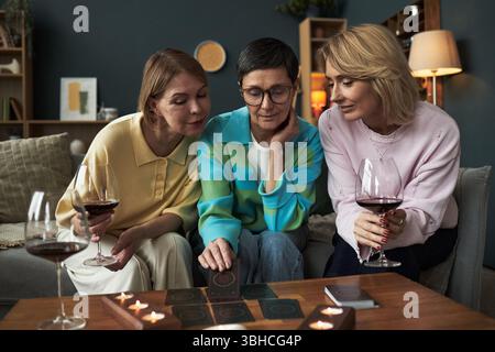 Trois femmes caucasiennes d'âge moyen assises ensemble sur un canapé tenant des verres de vin rouge, se livrant à la lecture de cartes de tarot avec des bougies allumées sur la table, souriant et interagissant étroitement Banque D'Images