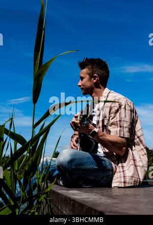 Un homme est assis sur un quai près de l'eau jouant de la guitare. Il porte une chemise à carreaux et un Jean, et a une montre sur son poignet gauche. Il regarde th Banque D'Images
