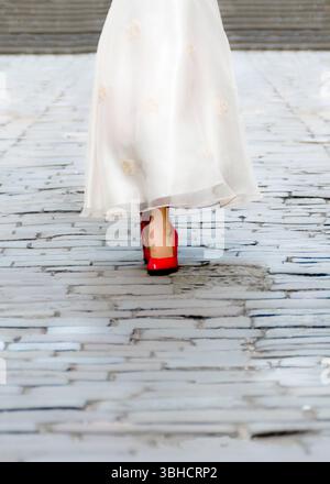 Une femme dans une longue robe blanche avec de petits détails de fleurs et des talons hauts rouge vif descend une passerelle en pierre grise. Banque D'Images