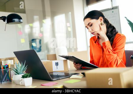 Jeune entrepreneure travaillant dans un bureau sur les ordres de livraison, concentrée sur un presse-papiers tout en gérant son entreprise de commerce électronique. Banque D'Images