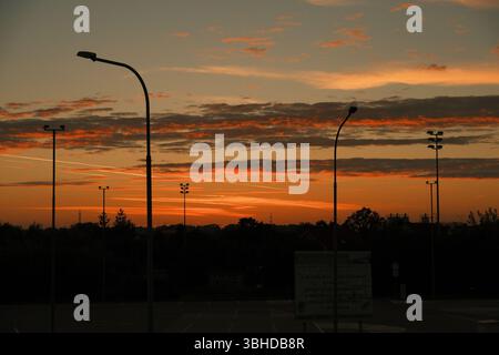 Scène de coucher de soleil urbaine avec des silhouettes de lampadaires et d'arbres sur un ciel dramatique peint avec des tons chauds orange et rouge. Banque D'Images