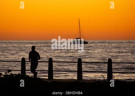 Silhouette d'un jogger au crépuscule sur la promenade du Cap, contre un ciel orangé doré avec un catamaran sur l'océan en arrière-plan. Banque D'Images