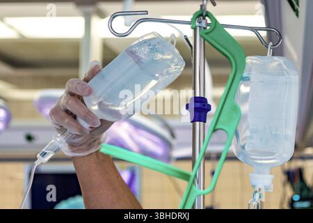 LK Osnabrück, Deutschland 30. Januar 2023 : Im Bild : dieses Bild zeigt eine hand einer medizinischen Fachkraft, die einen transparenten Einweghandschuh trägt und einen Infusionsbeutel mit einer klaren Flüssigkeit hält. Der Infusionsbeutel wird gerade an einem Infusionsständer befestigt oder davon abgenommen, was eine wichtige phase der Patientenversorgung im Krankenhaus darstellt. Ein zweiter Infusionsbeutel hängt bereits sicher am Ständer, was auf eine kontinuierliche Flüssigkeitszufuhr oder Medikamentenverabreichung hinweist. IM Hintergrund sind unscharfe medizinische Lampen mit ihrem charakt Banque D'Images