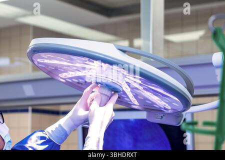 LK Osnabrück, Deutschland 30. Januar 2023 : Im Bild : dieses eindringliche Bild zeigt medizinische Fachkräfte-Hände, die in blauen, sterilen Einweghandschuhen stecken und eine hochmoderne Operationslampe präzise justieren oder halten. Die große, runde OP-lampe selbst strahlt ein charakteristisches violettes LED-Licht aus, welches für optimal die Ausleuchtung des Operationsfeldes unerlässlich ist. IM Hintergrund sind weitere Elemente eines OP-Saals oder Behandlungsraums erkennbar, wie helle Deckenbeleuchtung und unscharfe, technische medizinische Geräte oder Bildschirme, die die moderne Infrastr Banque D'Images