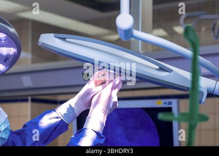 LK Osnabrück, Deutschland 30. Januar 2023 : Im Bild : dieses eindringliche Bild zeigt medizinische Fachkräfte-Hände, die in blauen, sterilen Einweghandschuhen stecken und eine hochmoderne Operationslampe präzise justieren oder halten. Die große, runde OP-lampe selbst strahlt ein charakteristisches violettes LED-Licht aus, welches für optimal die Ausleuchtung des Operationsfeldes unerlässlich ist. IM Hintergrund sind weitere Elemente eines OP-Saals oder Behandlungsraums erkennbar, wie helle Deckenbeleuchtung und unscharfe, technische medizinische Geräte oder Bildschirme, die die moderne Infrastr Banque D'Images