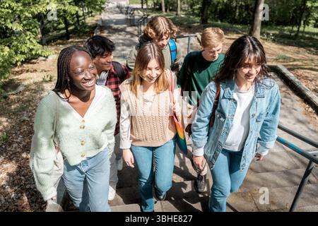 Groupe d'étudiants multiculturels qui montent les escaliers à l'extérieur dans un parc, souriant et parlant par une journée ensoleillée, symbolisant l'amitié et l'éducation Banque D'Images
