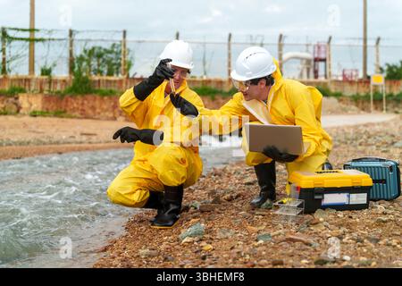 Deux scientifiques de l'environnement vêtus de combinaisons de protection jaunes et de casques recueillent des échantillons d'eau sur une rive tout en utilisant un ordinateur portable pour analyser les données sous Banque D'Images
