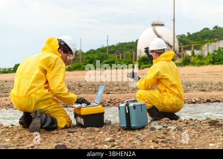 Deux scientifiques de l'environnement vêtus de combinaisons de protection jaunes s'agenouillent au bord de l'eau, à l'aide d'un ordinateur portable et d'outils pour analyser des échantillons lors d'une investig sur le terrain Banque D'Images