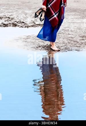 Une femme, portant un châle à carreaux rouge et une robe bleue, marche pieds nus le long du bord d'un plan d'eau peu profond. Elle porte ses talons hauts noirs en un seul Banque D'Images