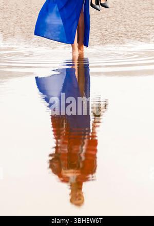 Une femme portant une longue robe bleue fluide marche pieds nus dans l'eau peu profonde, portant ses talons hauts noirs dans une main. Son reflet est visible i Banque D'Images