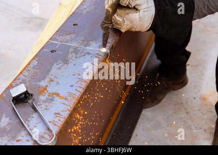 Travailleur qualifié coupe la poutre en acier, produisant des étincelles brillantes dans le chantier de construction en utilisant à travers Banque D'Images