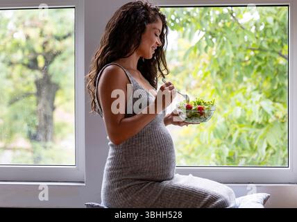 Photo confortable d'une femme avec un gros ventre enceinte assis près de la fenêtre de l'appartement pour manger sa salade Banque D'Images
