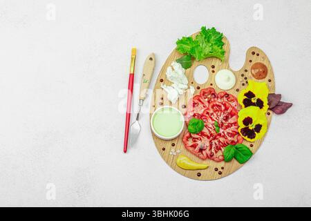 Tomate héritée tranchée avec feta, herbes, fleurs comestibles et sauces sur une palette d'artistes en bois, stylisée avec pinceau et couteau à palette. Concr. Pierre claire Banque D'Images