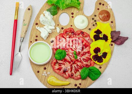 Tomate héritée tranchée avec feta, herbes, fleurs comestibles et sauces sur une palette d'artistes en bois, stylisée avec pinceau et couteau à palette. Concr. Pierre claire Banque D'Images