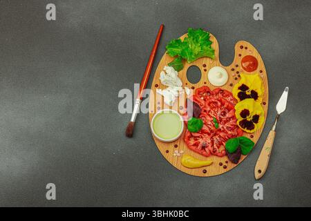 Tomate héritée tranchée avec feta, herbes, fleurs comestibles et sauces sur une palette d'artistes en bois, stylisée avec pinceau et couteau à palette. Concre de pierre sombre Banque D'Images