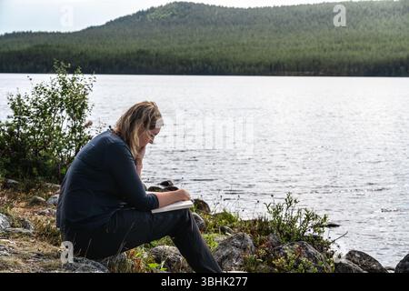 Une femme lit un livre alors qu'elle est assise au bord d'un lac par une brillante journée d'été en Suède. Elle profite de la paix et de la tranquillité de la nature. Banque D'Images