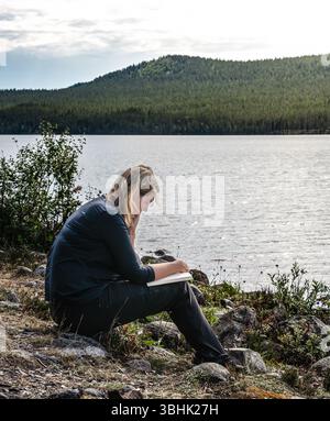 Une femme lit un livre alors qu'elle est assise au bord d'un lac par une brillante journée d'été en Suède. Elle profite de la paix et de la tranquillité de la nature. Banque D'Images