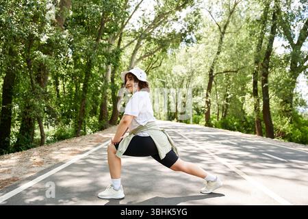 Une femme en forme s'étend le long d'une route tranquille entourée d'une végétation luxuriante tout en faisant de l'exercice. Banque D'Images