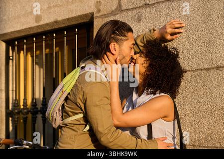 Jeune couple multi ethnique partageant un baiser dans une rue romantique de la ville, se prélasser dans la lueur chaude de la lumière du soleil heure d'or pendant l'été Banque D'Images