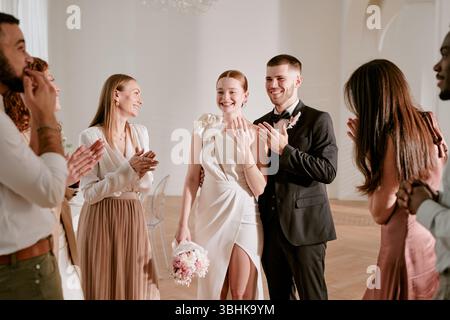 Jeune mariée et marié adultes caucasiens debout ensemble souriant tout en groupe multiethnique de jeunes amis adultes applaudissant et célébrant la cérémonie de mariage dans un cadre intérieur lumineux Banque D'Images