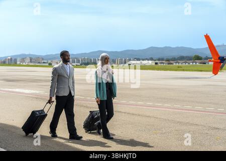 Homme d'affaires et femme d'affaires marchant le long du tarmac de l'aérodrome, tirant sur les bagages à main tout en profitant d'une journée ensoleillée de voyage et d'aventure Banque D'Images