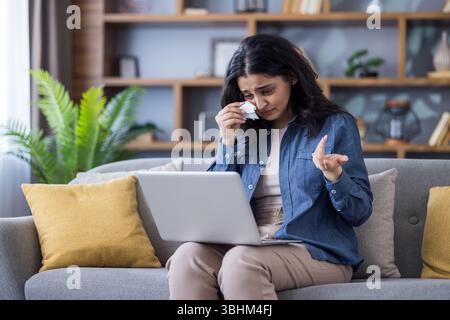 Bouleversée jeune femme indienne assise sur un canapé à la maison, tenant un ordinateur portable sur les genoux, parlant sur un appel vidéo et pleurant, essuyant les larmes avec une serviette. Banque D'Images