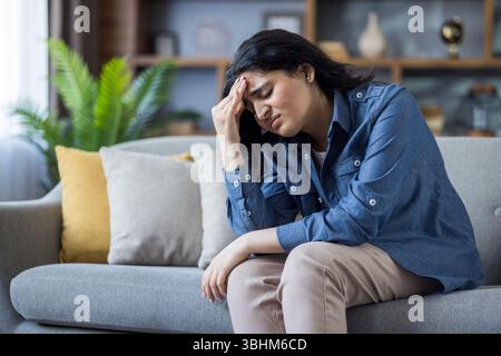 Une jeune femme éprouve un mal de tête alors qu'elle est assise sur un canapé à la maison, paraissant stressée. Elle se serre le front. Banque D'Images