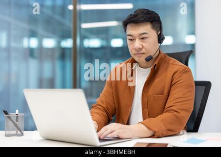 Un homme asiatique avec un casque utilise un ordinateur portable dans un environnement de bureau moderne, mettant en valeur le travail à distance et le service client. Banque D'Images