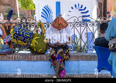 Chefchaouen, Chefchaouen - Maroc - 08-20-08 2024 ; homme en vêtements traditionnels avec un chapeau de paille décoré assis sur un mur de pierre, regardant son téléphone. Col Banque D'Images