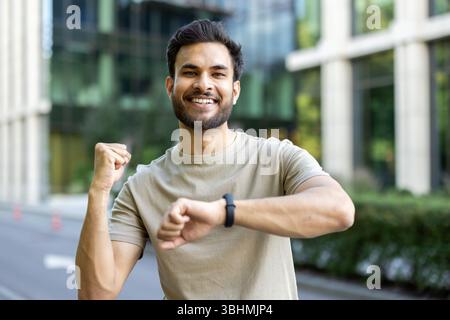 Portrait d'un jeune homme indien debout dehors, faisant du sport et de la course, regardant l'appareil photo avec joie et satisfaction du résultat sur la minuterie de la montre sur son poignet. Banque D'Images