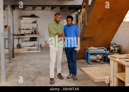 Collègues afro-américains discutant des plans en atelier avec tablette, générateur et panneaux de bois Banque D'Images