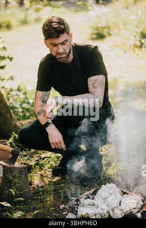 Un homme avec des tatouages sur les bras est assis dans l'herbe à côté d'un feu. Il est perdu dans ses pensées Banque D'Images
