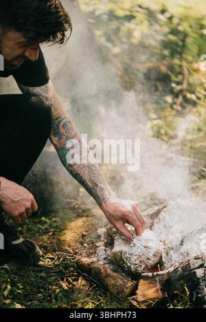 Un homme avec des tatouages sur le bras cuit de la nourriture sur un feu. L'homme sourit en préparant la nourriture Banque D'Images