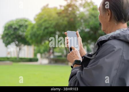 Photo d'un homme barbu senior asiatique portant un manteau de sport debout à l'extérieur au parc au coucher du soleil à l'aide d'un téléphone portable. Copier l'espace. Banque D'Images