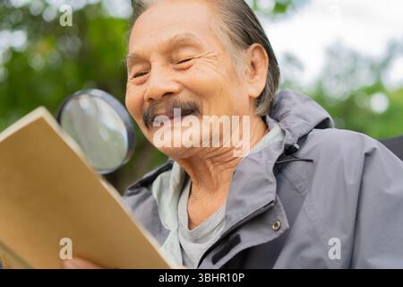 Vieil homme aîné asiatique myope , myope , lisant un livre avec une loupe en plein air au parc - homme senior de style de vie ont la prévoyance pro Banque D'Images