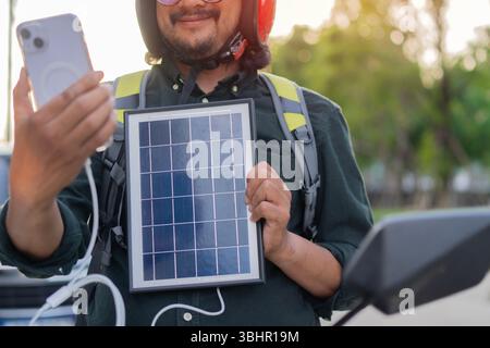 Homme barbu heureux assis sur une moto chargeant des gadgets avec panneau solaire portable, technologie moderne pour le voyage. Source d'électricité alternative Banque D'Images