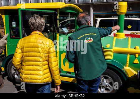 Femme en veste jaune se tient devant le mini train touristique de Cologne et a quelque chose expliqué par le chauffeur, Cologne, Allemagne. Frau in gelb Banque D'Images