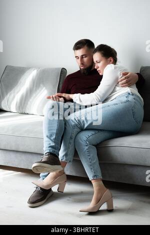 Un couple profite d'un moment tendre sur un canapé gris moelleux, avec une lumière naturelle douce illuminant leur connexion intime alors qu'ils se câlinent étroitement pendant un p Banque D'Images