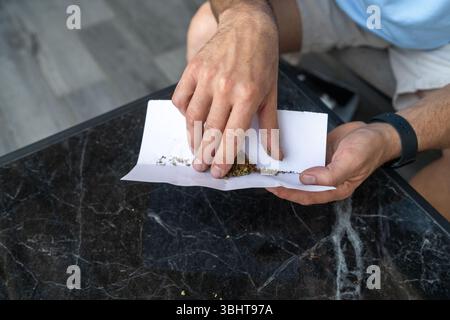 Une personne prépare habilement un joint de cannabis, répandant méticuleusement du cannabis moulu sur du papier à rouler. Le cadre dispose d'une table en marbre élégante, reflétant une ambiance décontractée l'après-midi. Banque D'Images