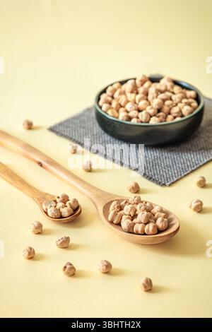 Pois chiches séchés sains dans des cuillères en bois et bol en céramique sur fond pastel. Arrangement de nourriture rustique avec lumière naturelle Banque D'Images