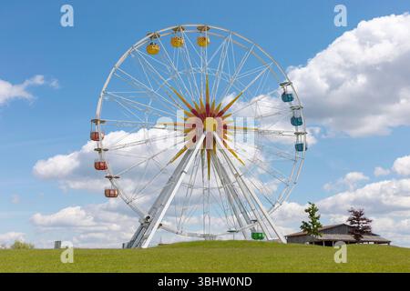 Une grande roue avec des gondoles colorées est exposée bien en évidence contre un ciel bleu éclatant rempli de nuages moelleux. L'herbe environnante est Banque D'Images