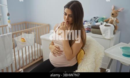 Femme assise dans la chambre de bébé sur la chaise enceinte réfléchie tenant le ventre contemplant la future pépinière Banque D'Images