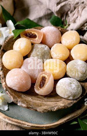Gâteaux de riz mochi japonais traditionnels colorés avec une garniture sucrée sur une assiette en bois rustique décorée de fleurs blanches et de feuilles vertes Banque D'Images