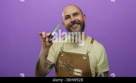 Homme barbu chauve portant un tablier et tenant des ciseaux sourit en toute confiance sur un fond violet vif mettant en vedette des bras tatoués et la coiffure t Banque D'Images