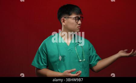 Jeune homme dans des gommages verts sur un fond rouge vif gestes avec les deux mains comme si vous présentiez quelque chose, transmettant un professionnel médical curieux s Banque D'Images