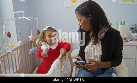 Mère utilisant le téléphone pendant que la fille joue à côté d'un berceau dans une chambre confortable, soulignant l'amour de la famille et la connexion. Banque D'Images