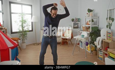 Homme dansant joyeusement dans une crèche colorée remplie de jouets et de meubles, mettant en valeur une atmosphère intérieure heureuse et animée. Banque D'Images