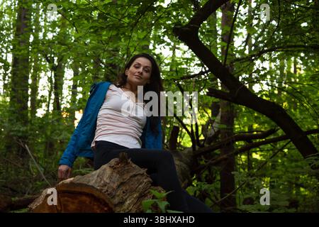 Femme de beauté posant dans un manteau bleu de mode, regardant devant la forêt de pins. Femelle se détendre assis dans un parc sur une vieille bûche pourrie. Fille pensant et ressentant Banque D'Images
