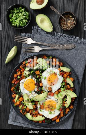 Cubes de patates douces frits et servis avec des œufs ensoleillés et des tranches d'avocat frais sur une assiette sur une table en bois noir avec fourchettes et serviette, à la verticale Banque D'Images