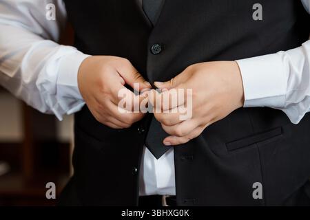 Un gros plan d'un beau marié attachant les boutons de son gilet noir sur une chemise blanche bien nette tout en préparant sa cérémonie de mariage Banque D'Images